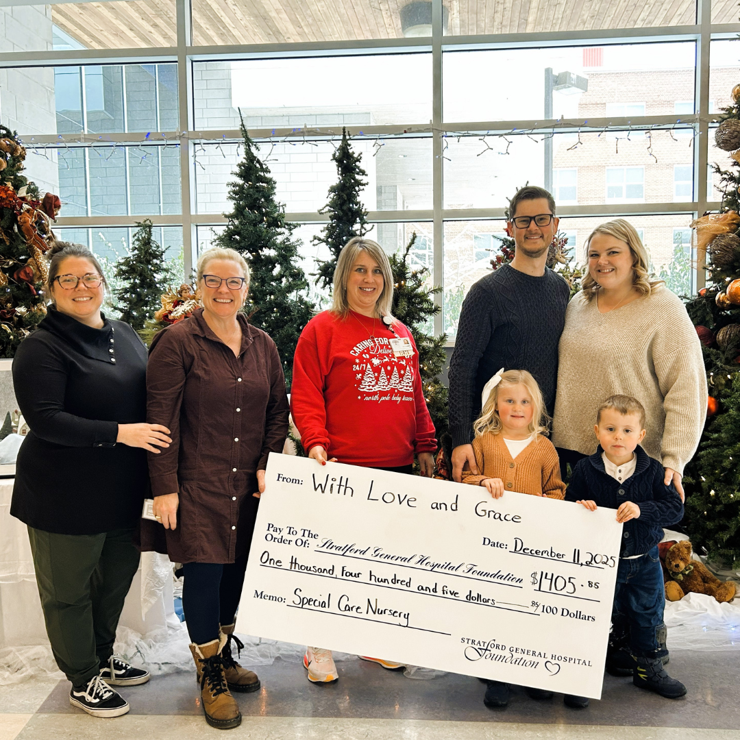 Five adults and two young children stand in front of decorated Christmas trees inside Stratford General Hospital, holding a large ceremonial cheque from With Love and Grace for $1,405.85 in support of the Special Care Nursery.